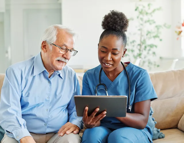 immigration-physicals-img Healthcare professional shows tablet to an elderly man at home while sitting on a couch.