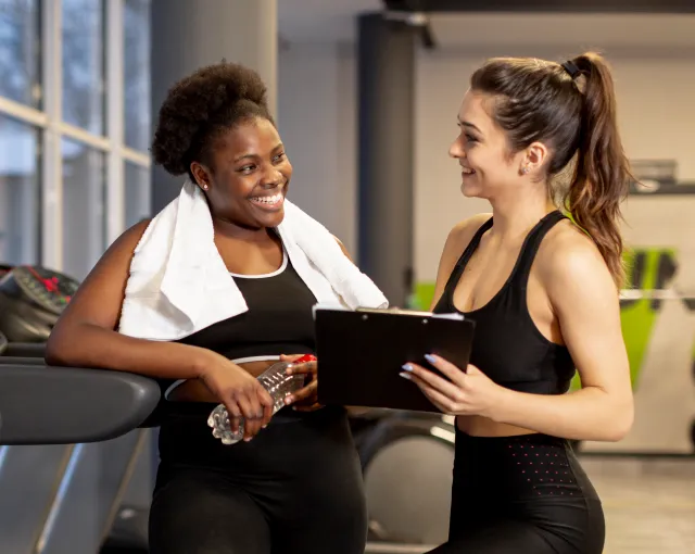 optional-services-img Two women in a gym chatting; one holds a water bottle with a towel draped over her shoulders, the other holds a clipboard.