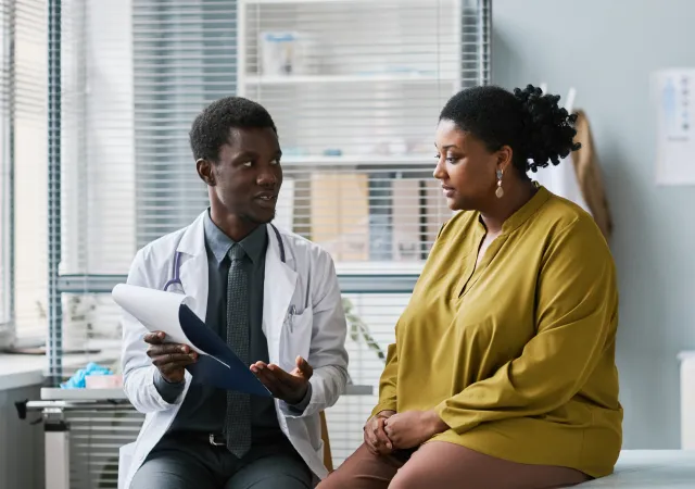 patient-info-img Doctor in a white coat showing a clipboard to a seated patient in a clinic, discussing documents.