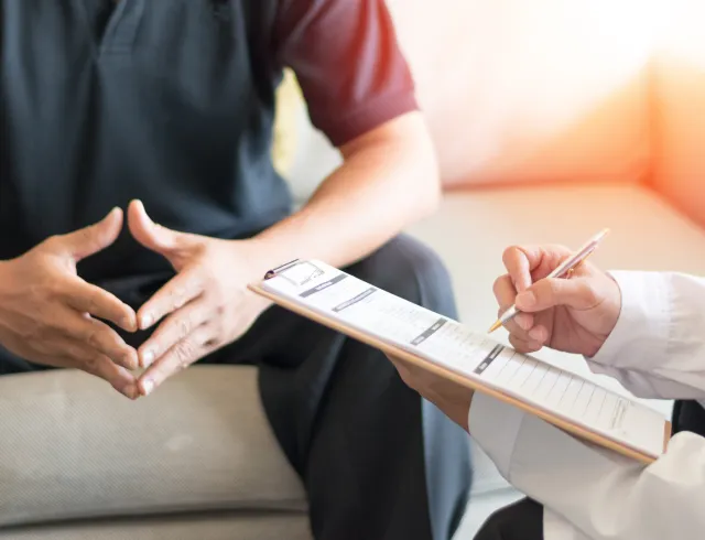 personal-injury-services-img Patient sits on a sofa with hands resting together while clinician writes on a clipboard nearby, documenting the session.